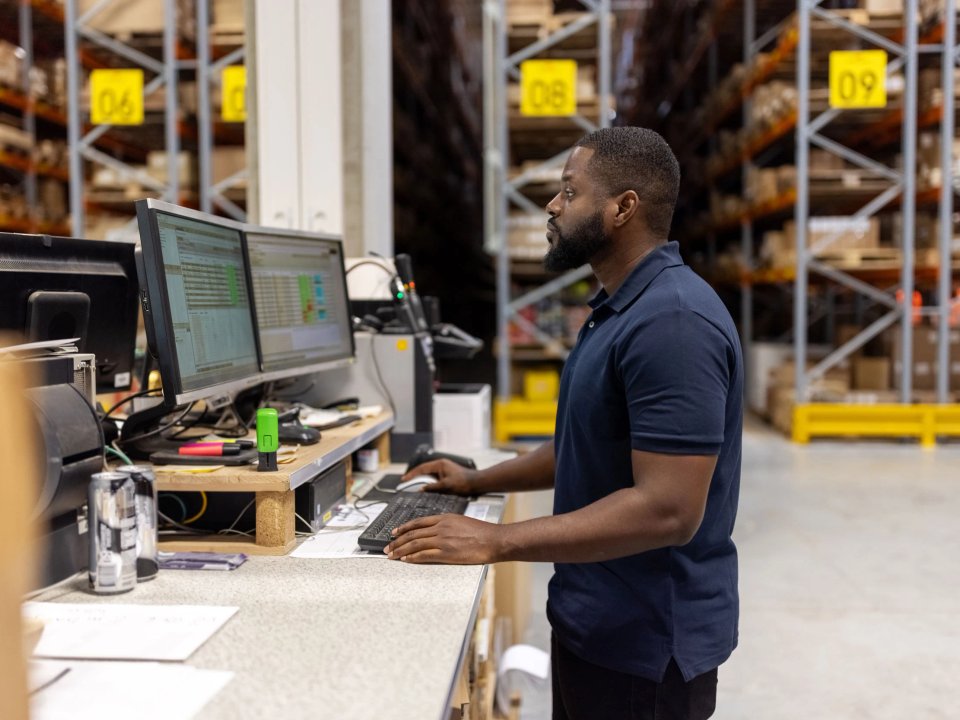 Warehouse team member reviewing inventory data on dual monitors at a workstation, with numbered pallet rack labels visible behind him