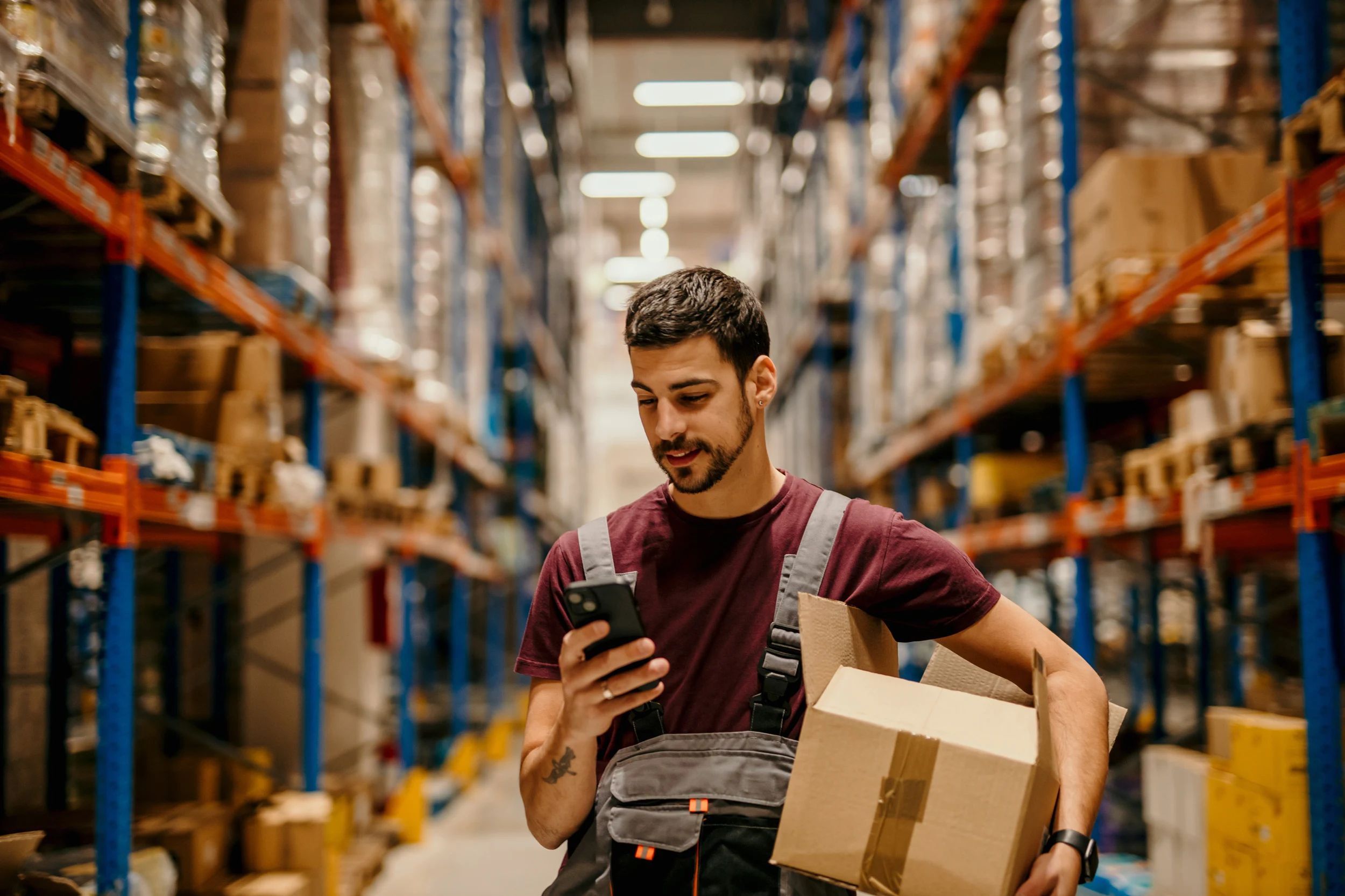 Warehouse worker checking his phone while carrying a carton through an aisle